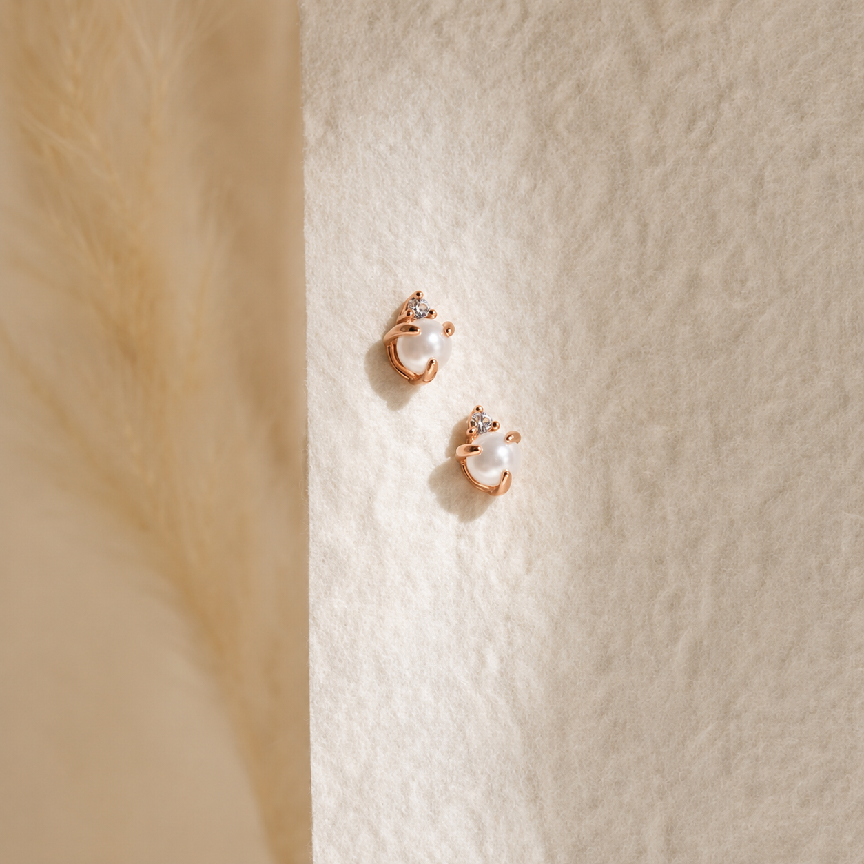 A pair of small rose gold huggie hoop earrings with white pearl centers and tiny clear crystal accents, displayed on a textured cream surface.
